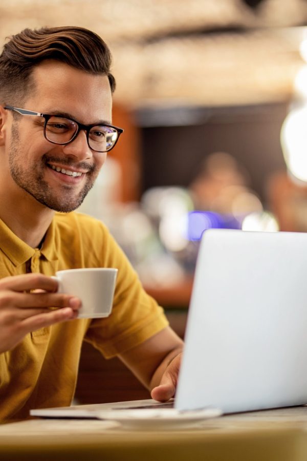 Smiling man surfing the net on laptop while drinking coffee in a bar.